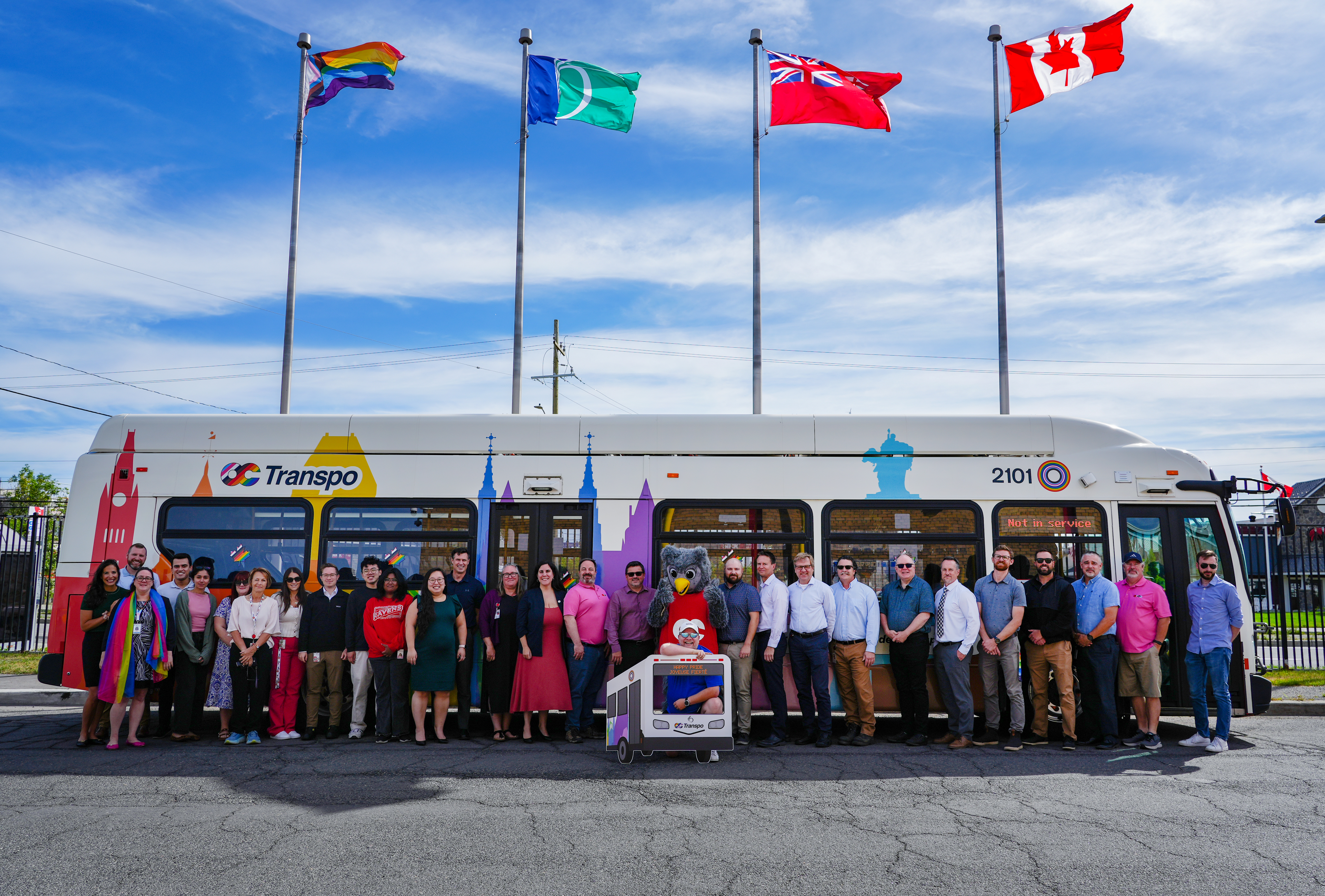 Un groupe d’employés d’OC Transpo pose devant un autobus blanc avec un habillage aux couleurs de la Fierté, avec des drapeaux qui flottent au-dessus.
