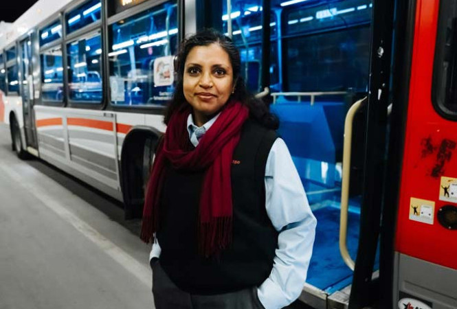 A woman bus driver stands in uniform in front of the side door of the bus