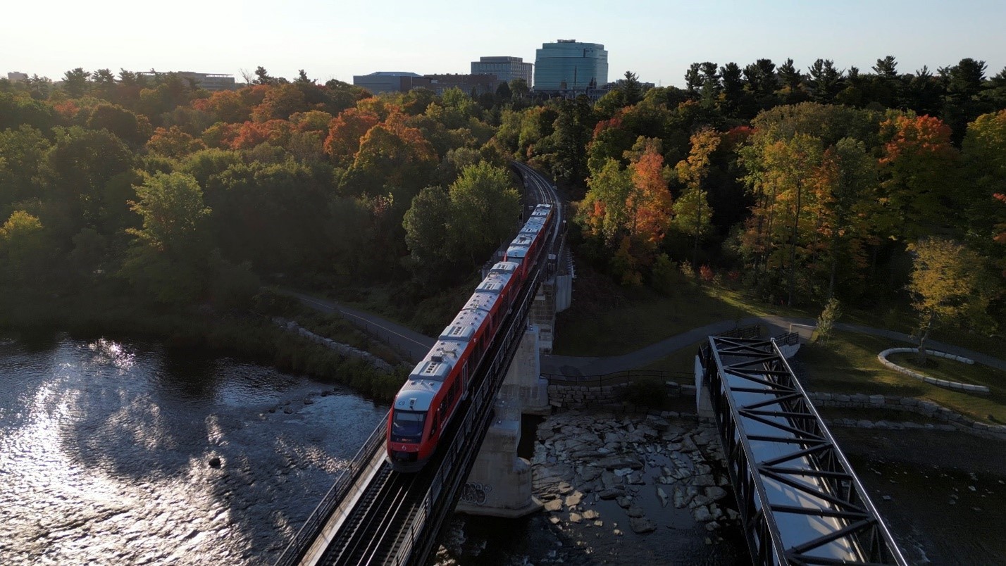 A Stadler FLIRT train crossing the bridge next to Carleton University over the Rideau River, with autumn trees and city buildings in the background.
